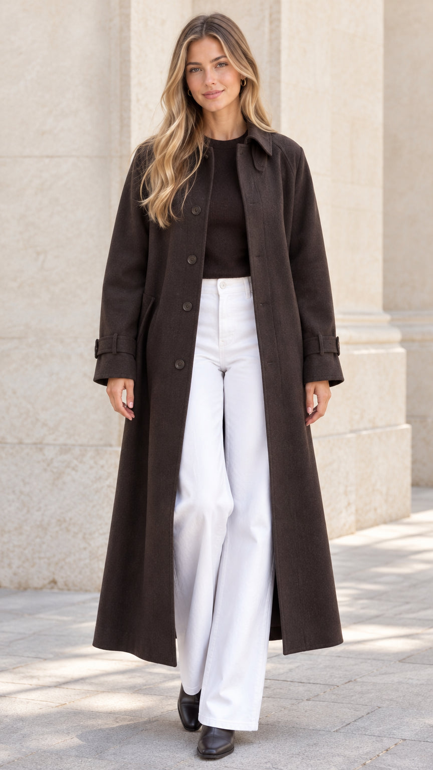 Front full view of woman wearing a dark brown full-length overcoat with white wide-leg trousers