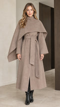 Front view of woman wearing taupe capelet overlay belted long overcoat with black boots