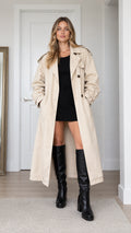 Full front view of model wearing beige long double-breasted trench coat with black dress and high boots