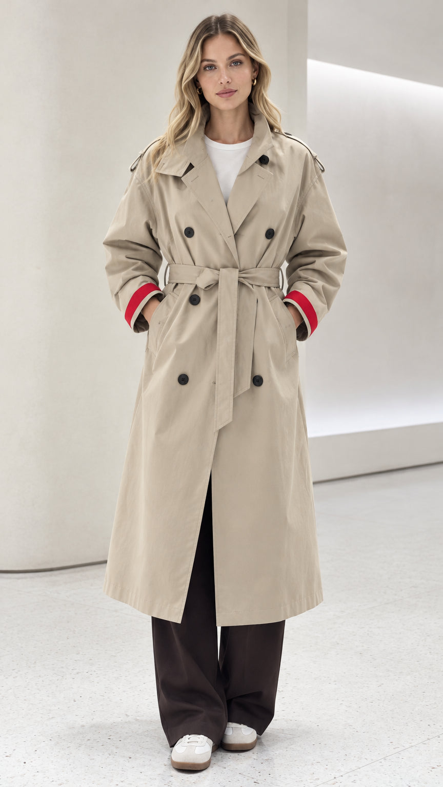 Front view of model wearing beige double-breasted long trench coat with red contrast cuffs and tied belt