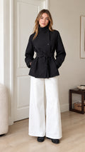 Front view of model wearing black funnel neck belted short trench coat with white wide-leg trousers