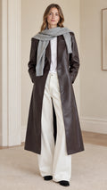 Front view of model wearing dark brown longline faux leather trench coat paired with white trousers and grey scarf