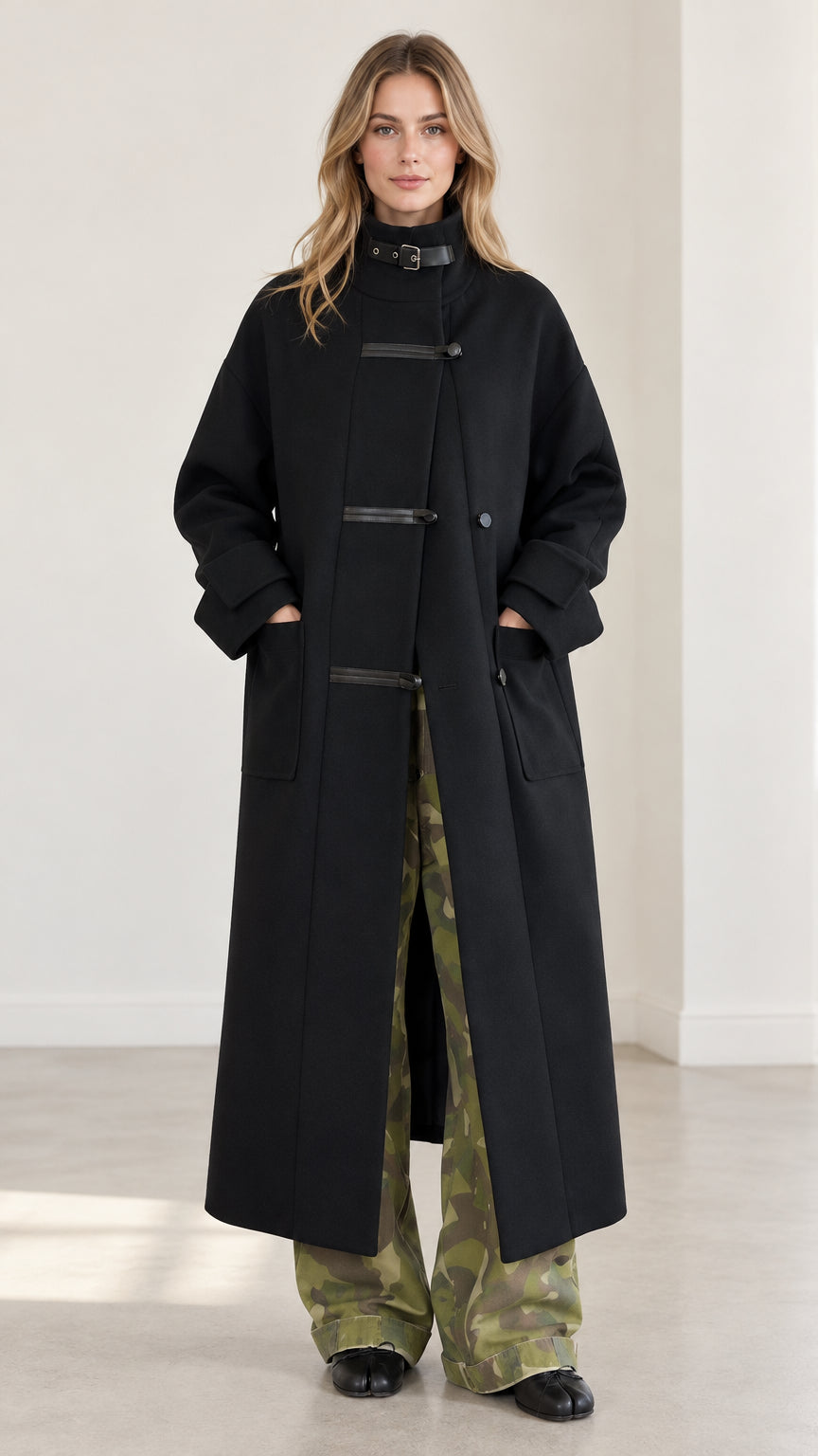 Front view of model wearing full-length black high-neck overcoat with buckle collar and camo trousers