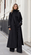 Front full-length view of woman wearing black maxi tailored overcoat with contrast collar and matching wide-leg trousers