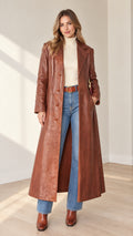 Front view of woman wearing full-length brown faux leather trench coat over cream turtleneck and flared jeans