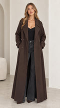 Full front view of model wearing a floor-length brown double-breasted trench coat with dark trousers