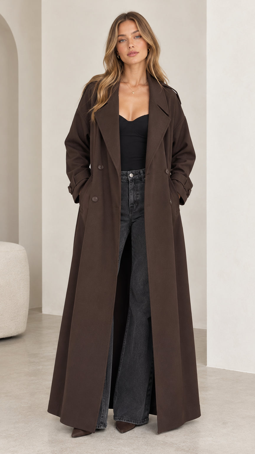 Full front view of model wearing a floor-length brown double-breasted trench coat with dark trousers