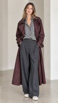 Model wearing long burgundy belted trench coat with open front over plaid shirt and trousers