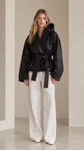 Front view of model wearing black hooded belted faux leather wrap jacket with white wide leg pants