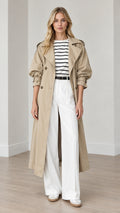 Full-length front view of woman wearing khaki double-breasted long trench coat with striped top and white trousers