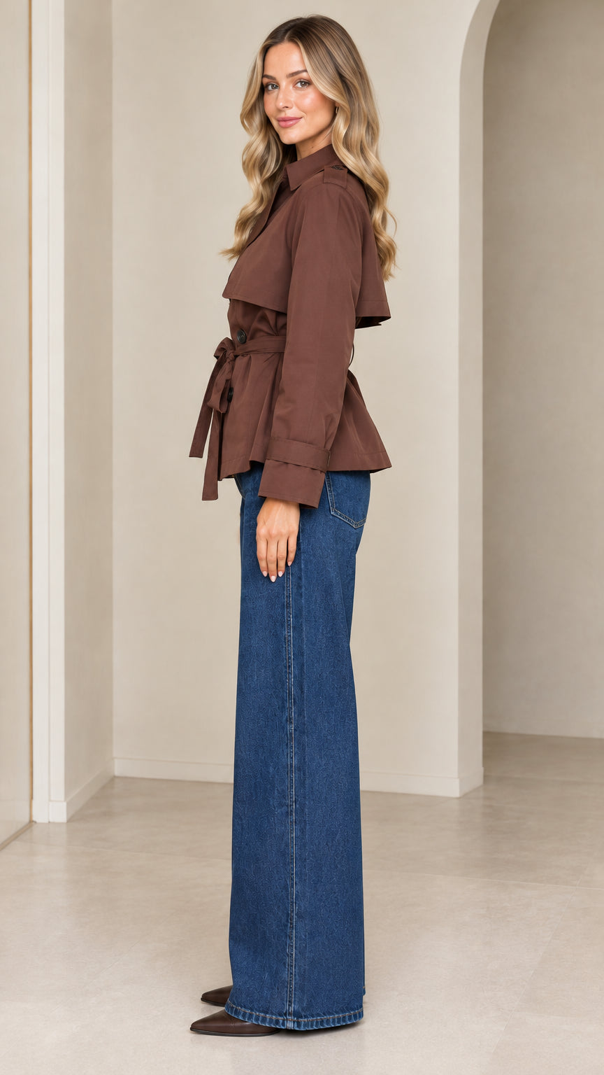 Side profile view of model wearing a brown cropped belted trench coat paired with denim trousers