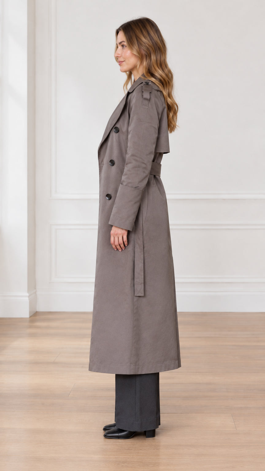 Side profile view of woman wearing taupe long trench coat with waist belt