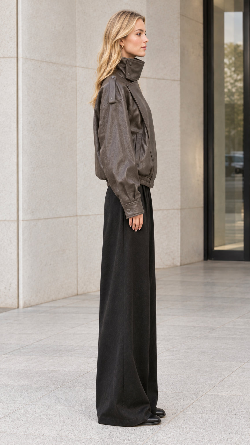 Side full-body view of model wearing charcoal brown faux leather jacket paired with black trousers