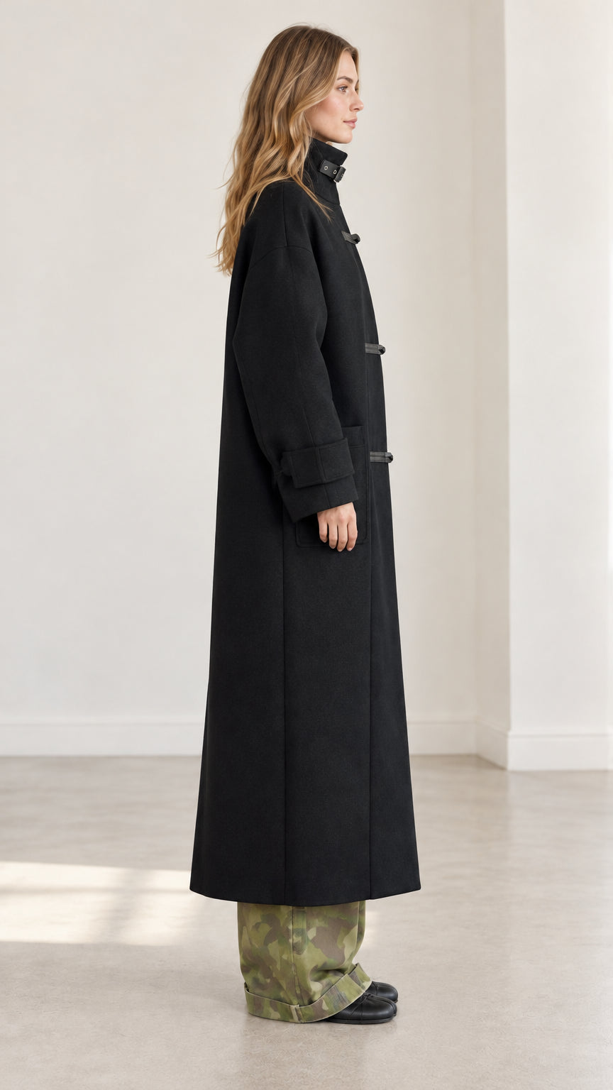 Side profile view of woman wearing black longline overcoat with stand collar and dropped shoulders