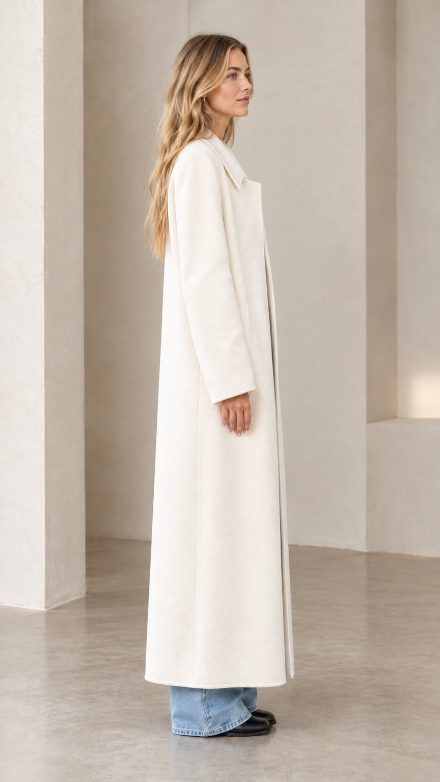 Side profile view of a model showcasing the straight silhouette of the cream floor-length overcoat