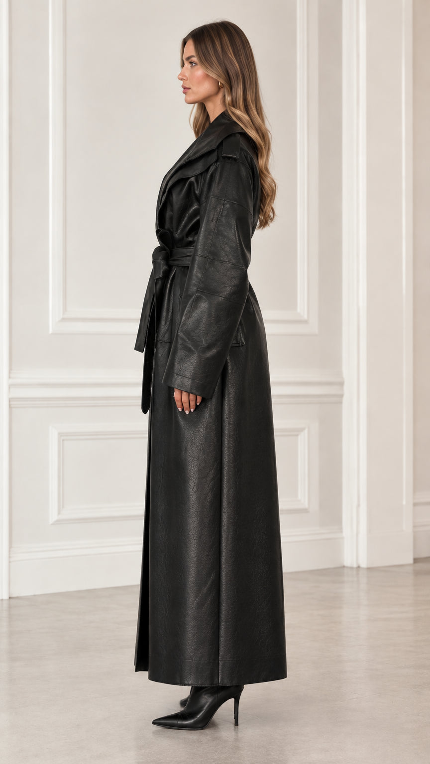Side profile of model in full-length black faux leather belted trench coat