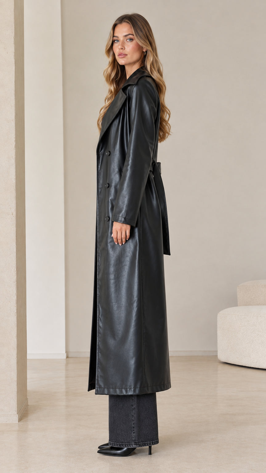 Side profile of model wearing a long black faux leather trench coat showing tailored silhouette and length