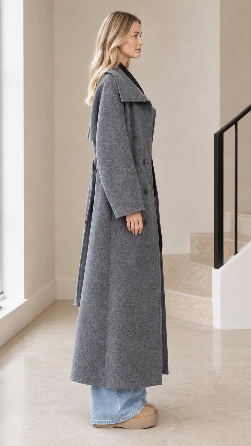 Side profile view of model wearing grey full-length belted overcoat
