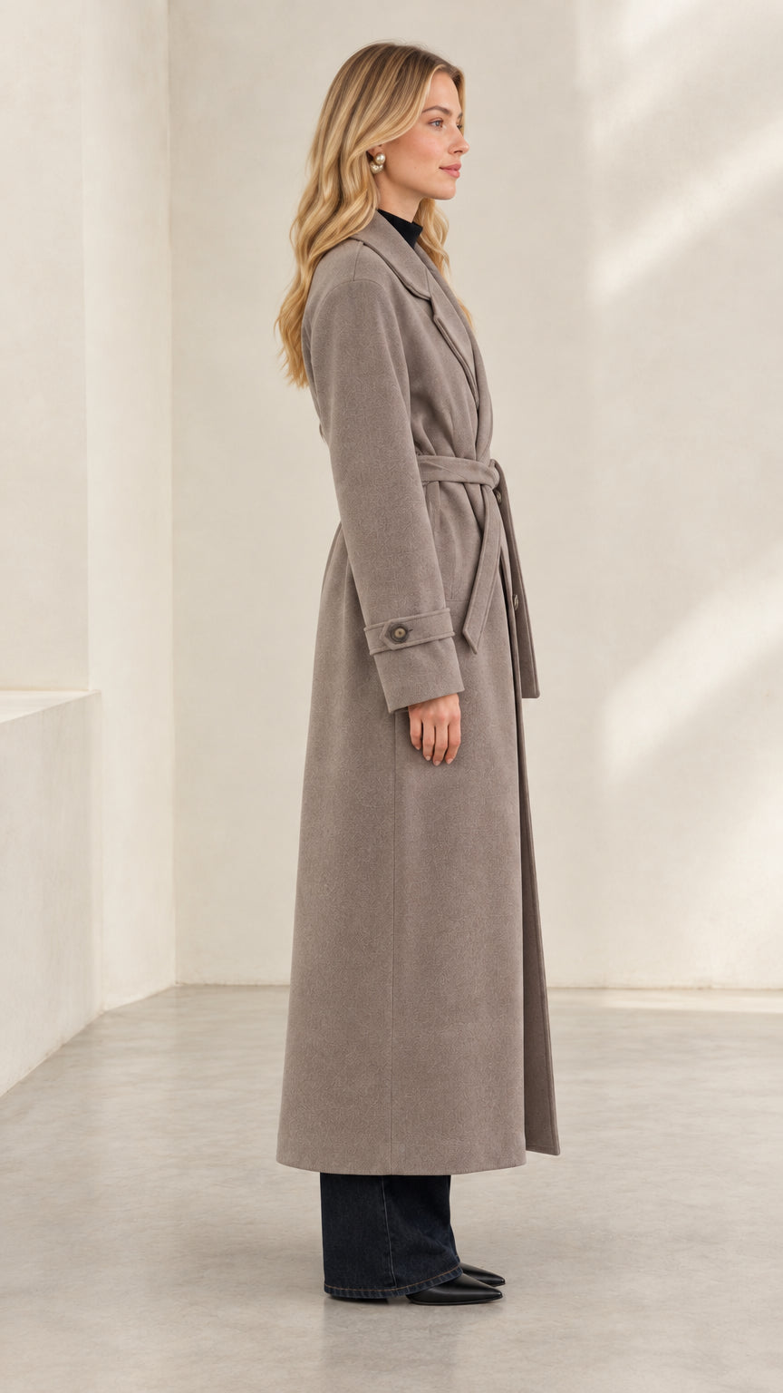 Side profile view of woman wearing taupe long overcoat showing maxi length and sleeve cuff tab