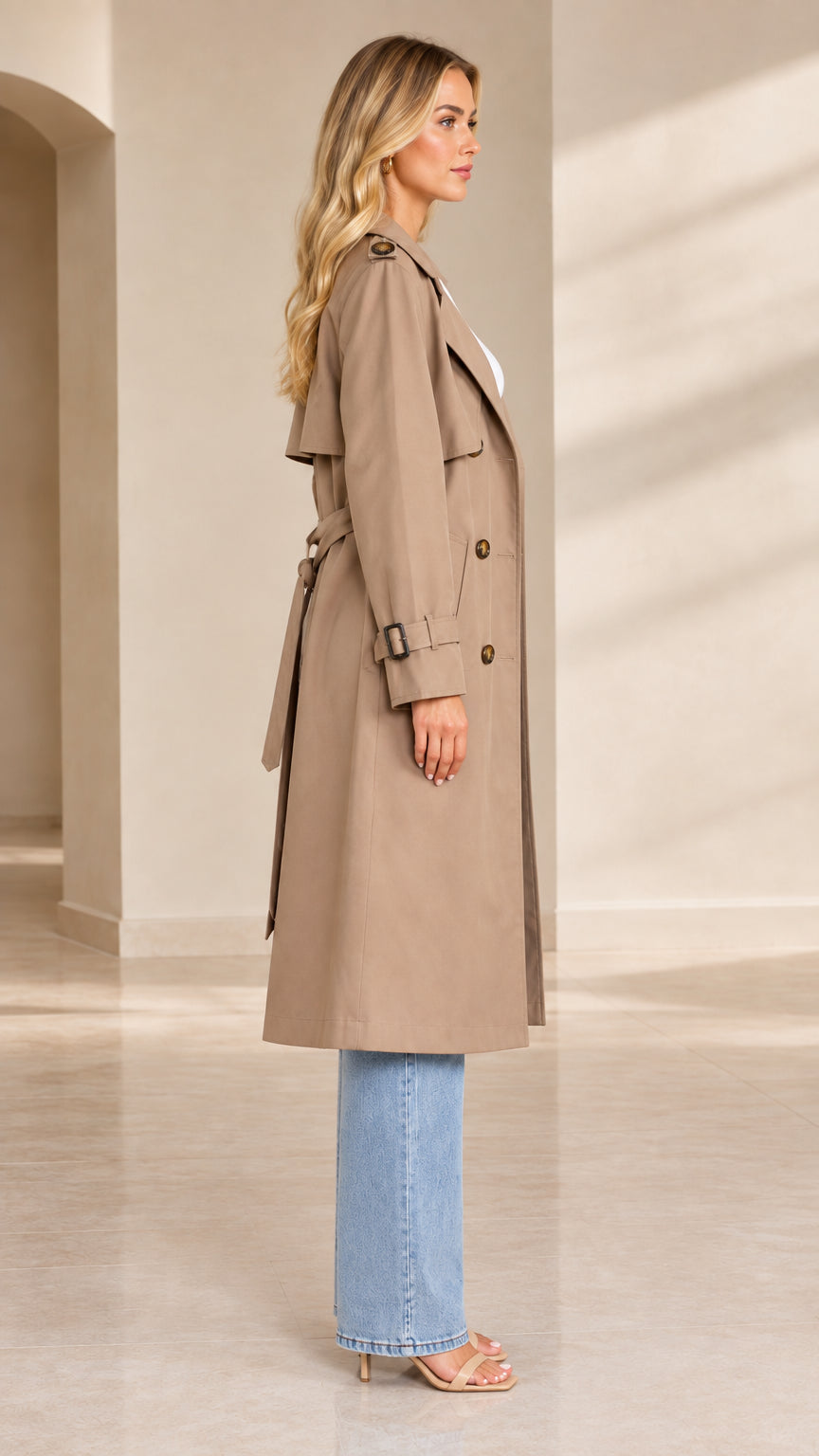Side profile of model wearing khaki double-breasted trench coat with waist belt
