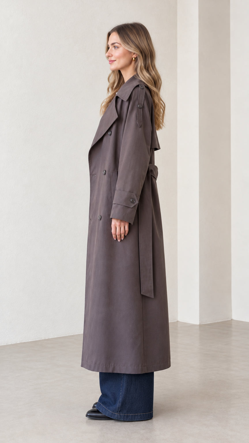 Side profile of woman modeling ankle-length charcoal trench coat with tie belt