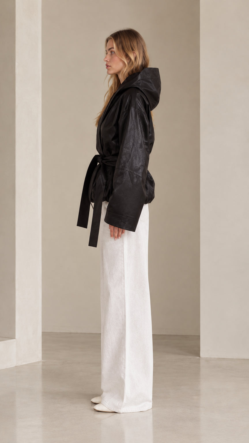 Side profile view of black hooded faux leather wrap jacket showing wide sleeves and tie belt