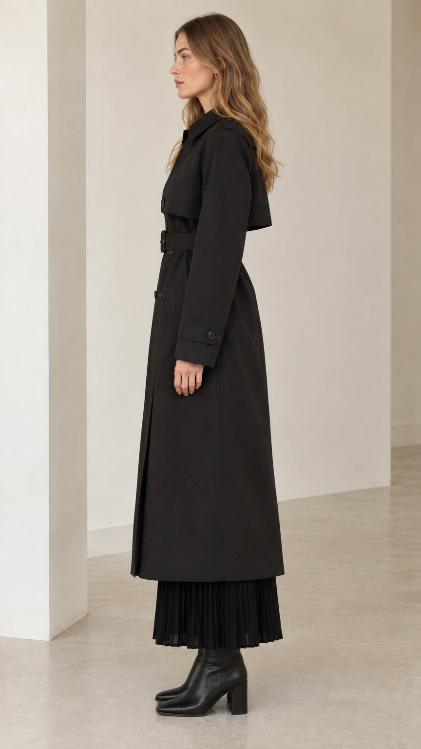 Side profile of model wearing full-length black trench coat with waist belt
