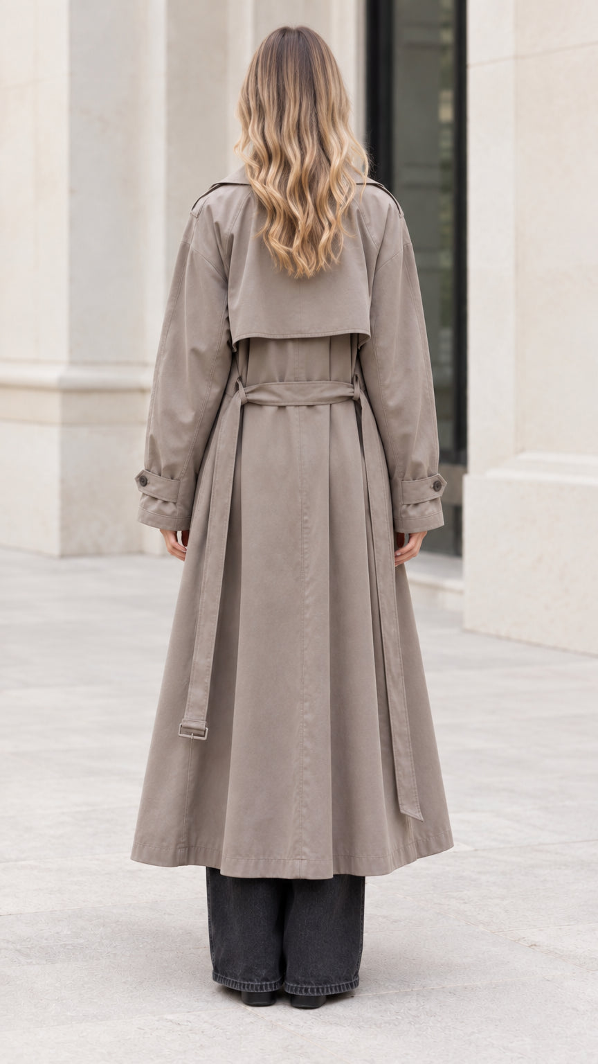 Back view of woman wearing taupe longline trench coat showing storm flap and belt