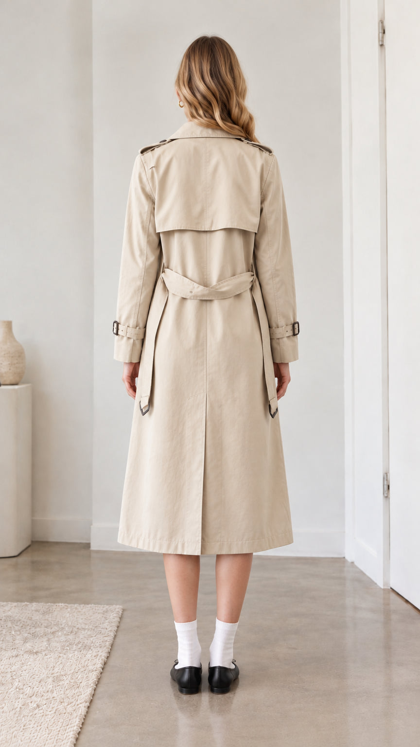 Back view of model wearing belted beige long trench coat showing storm flap detail