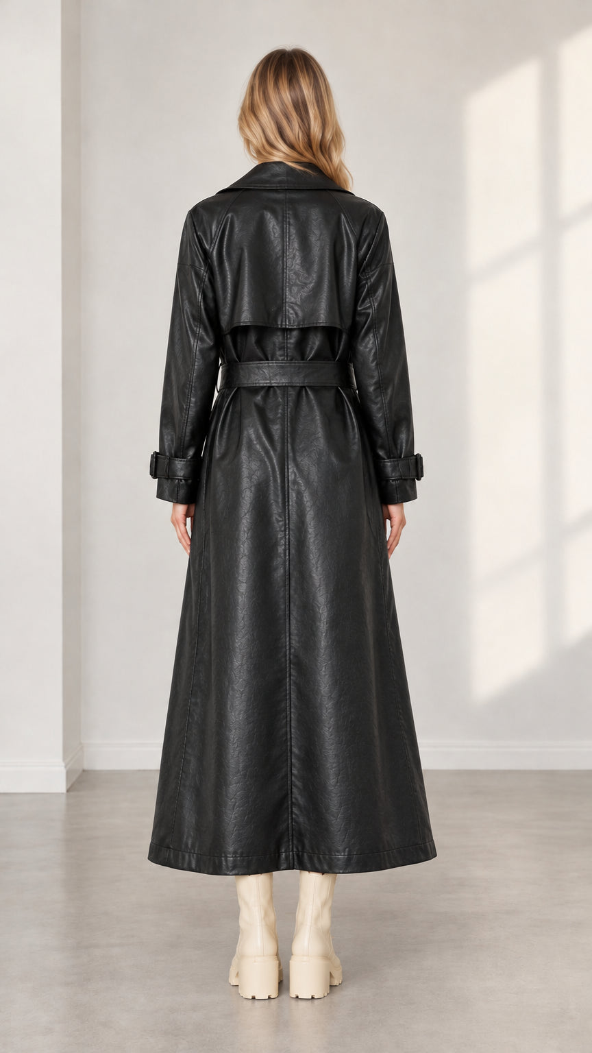 Back view of woman wearing belted black faux leather maxi trench coat showing storm flap
