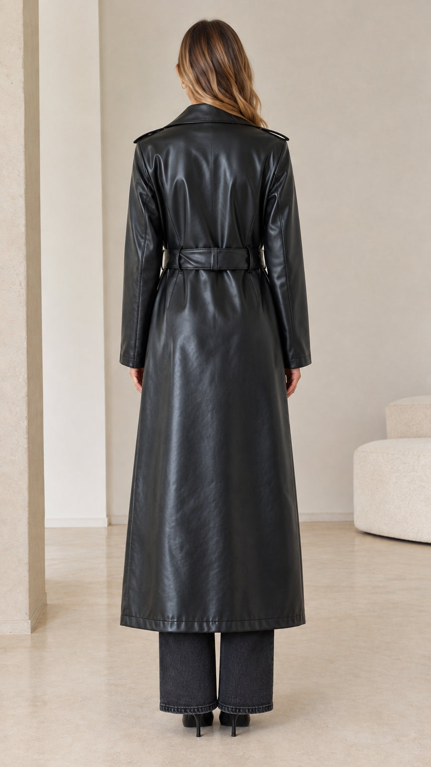 Rear view of model wearing a black faux leather maxi trench coat cinched at the waist with a buckled belt