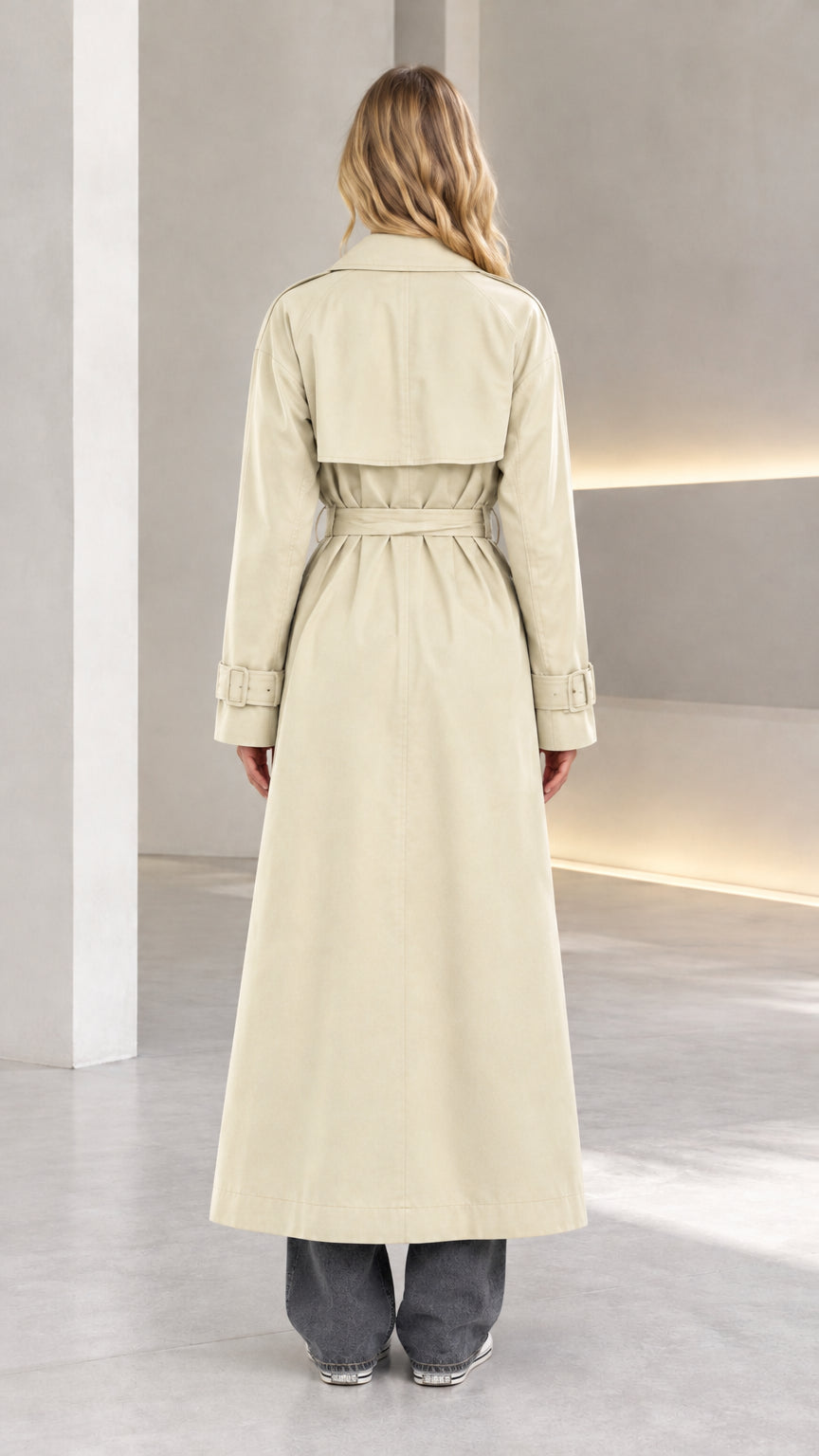 Back view of model wearing beige long trench coat highlighting storm flap and waist belt