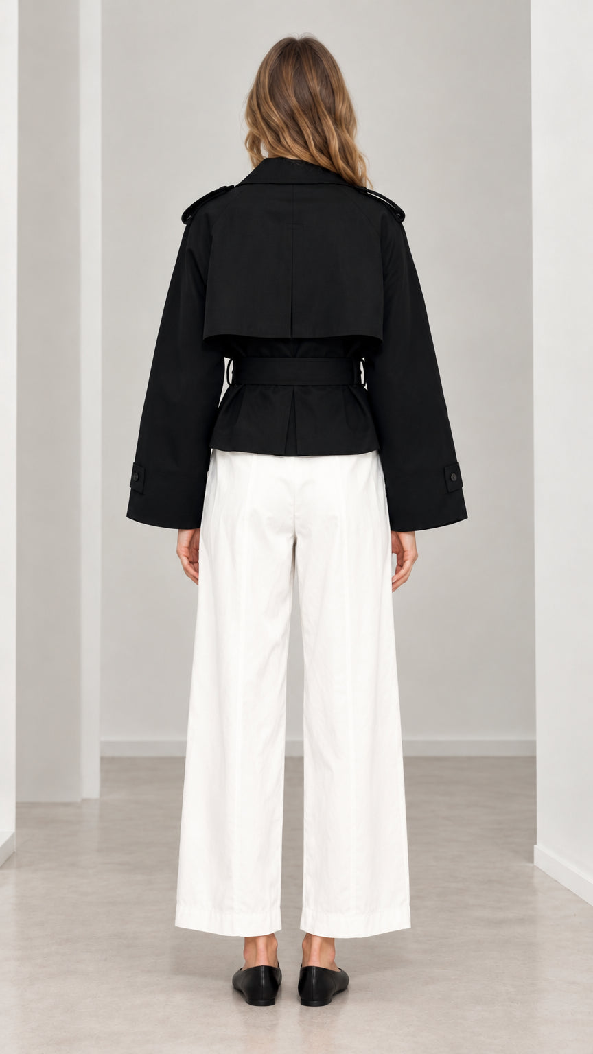 Back view of woman wearing black cropped trench jacket showing back storm flap and cinched belt