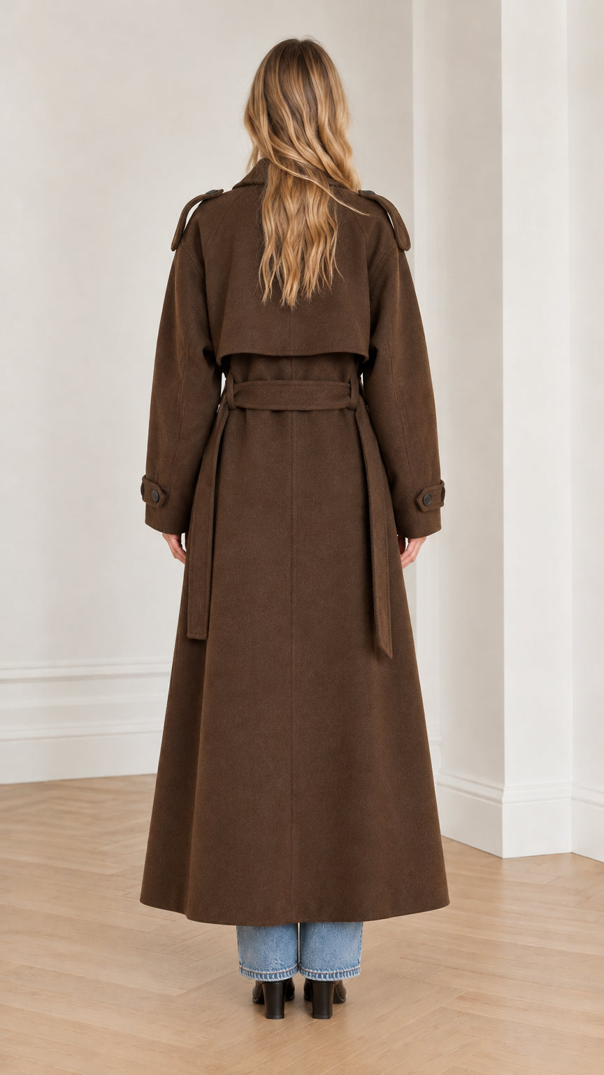 Back view of woman wearing brown maxi trench coat featuring back storm flap and tie belt