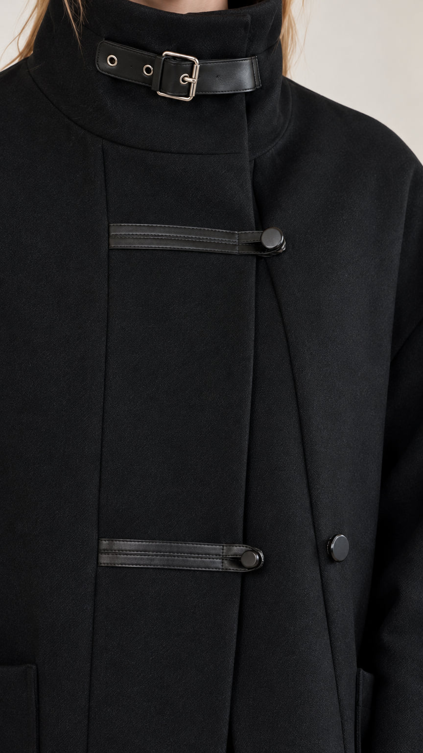 Close-up detail of high collar buckle closure and contrast black tab button placket on black overcoat