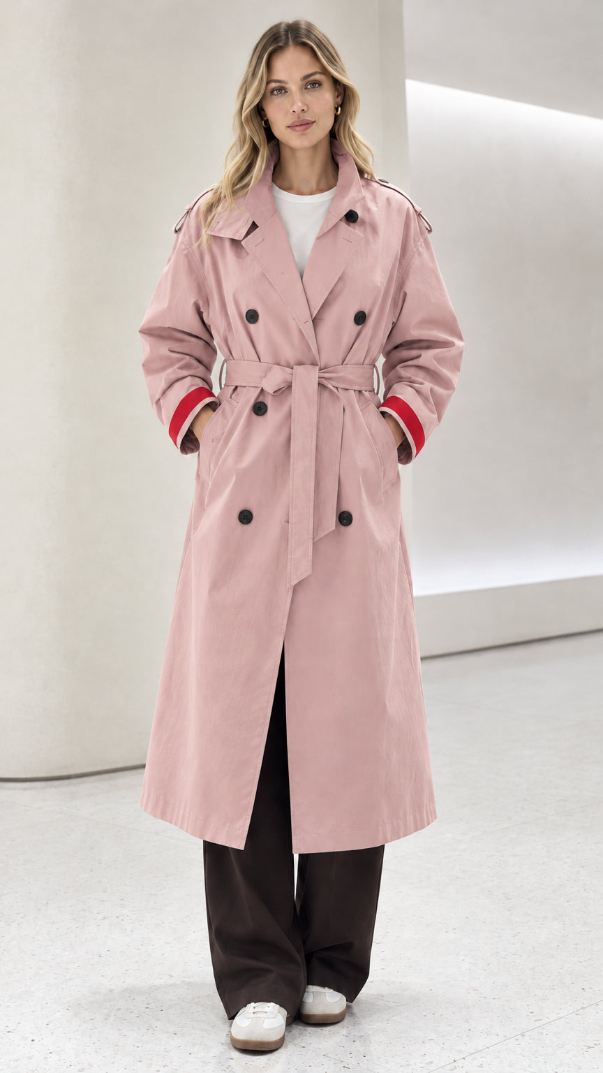 Front view of model wearing pastel pink double-breasted belted trench coat with red cuffs